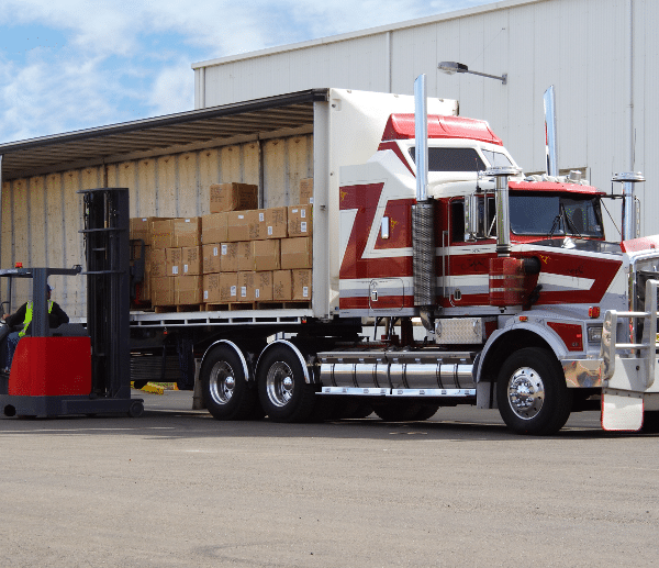 Forklift loading boxes into a red and white semi-trailer truck at a warehouse facility, illustrating less-than-truckload (LTL) freight shipping.