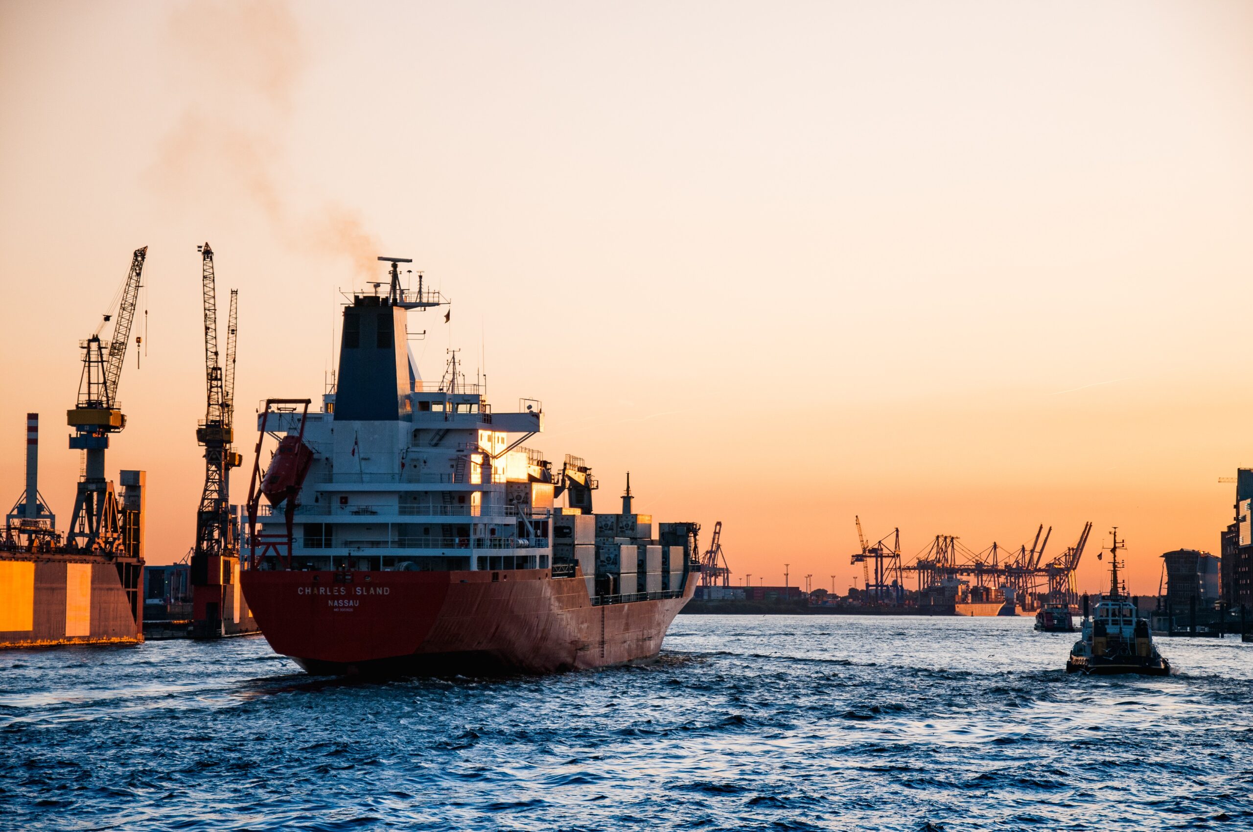 Large red cargo ship sailing into a busy industrial port at sunset, with dock cranes and containers in the background under an orange sky.